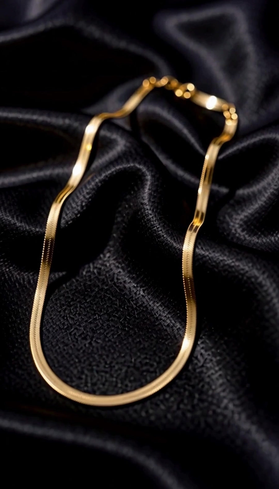 The Signature Chain, a gold herringbone chain, on black satin
